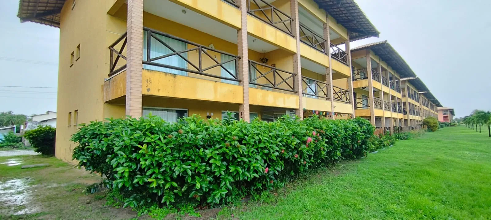 Bloco amarelo do Villa das Águas com arbustos floridos e gramado com coqueiros ao fundo